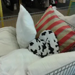 Wózek sklepowy wypełniony różnorodnymi poduszkami: białe, w paski i z motywem krowy, tworzące przytulną i eklektyczną kompozycję.