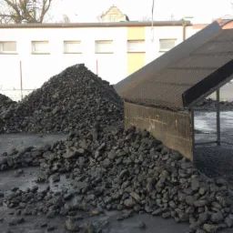 Sterta ekogroszku na placu składowym obok metalowej konstrukcji przesiewacza. W tle biały budynek z rzędem okien i drzewa bez liści.