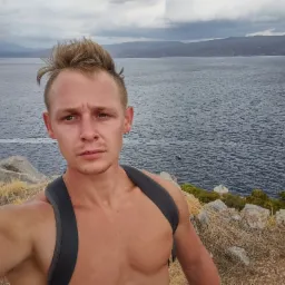 Młody mężczyzna robiący selfie na tle morza i górzystego krajobrazu pod pochmurnym niebem, z widoczną łodzią na wodzie.