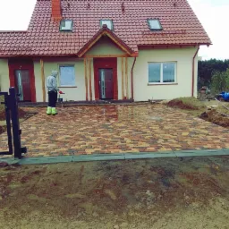 Nowo ułożona, wielokolorowa kostka brukowa na podjeździe przed domem, myta pod ciśnieniem przez pracownika w odblaskowej odzieży. Widoczne przygotowane obrzeża i brama wjazdowa.