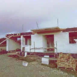 Budowa domu jednorodzinnego z białych bloczków, widoczne rusztowanie, częściowo wykonana konstrukcja dachu, cegły na palecie, teren budowy zarośnięty trawą.