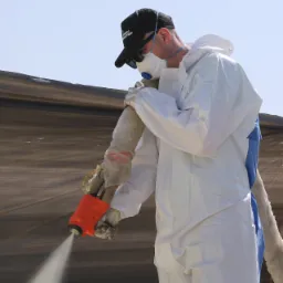 Pracownik w kombinezonie ochronnym i masce natryskuje izolację na ciemną powierzchnię, prawdopodobnie dach, używając specjalistycznego pistoletu natryskowego z pomarańczową osłoną.
