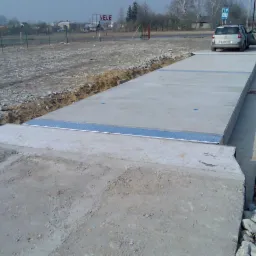 Betonowa waga samochodowa z najazdem, srebrnym wykończeniem i zaparkowanym obok autem z otwartymi drzwiami na placu budowy.