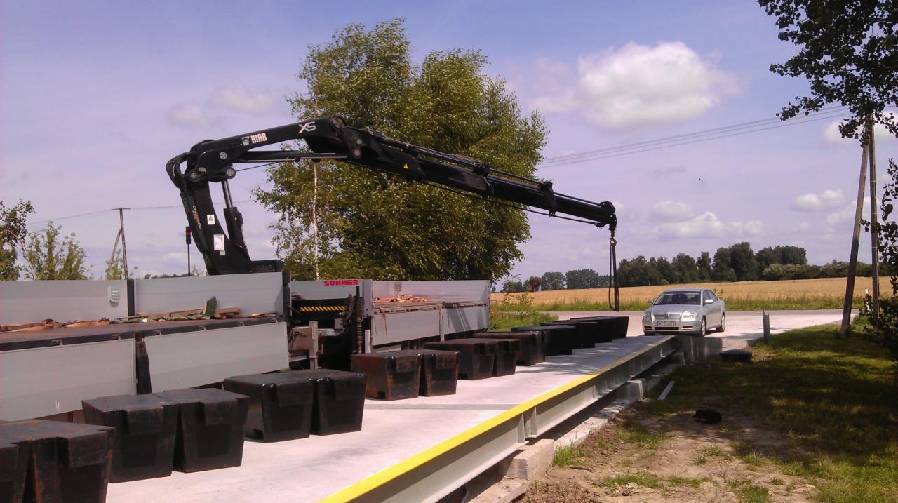 Ciężarówka z dźwigiem Hiab XS, przygotowana do transportu elementów, z samochodem osobowym w tle, na tle pola i błękitnego nieba.