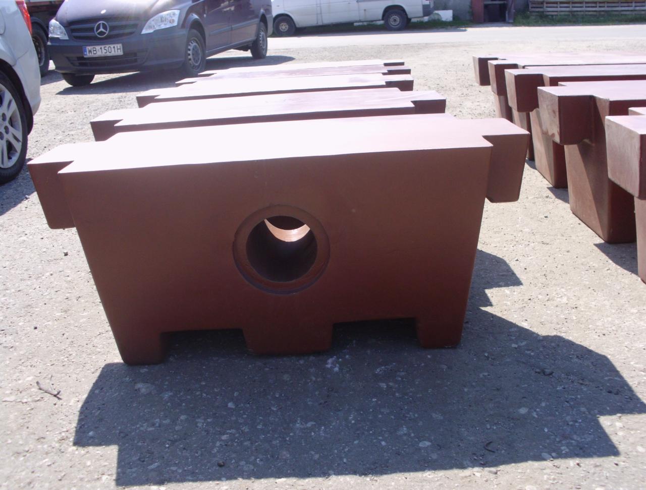 Ciemnobrązowe, metalowe elementy maszyn z okrągłym otworem na betonowym podłożu. W tle zaparkowane pojazdy.