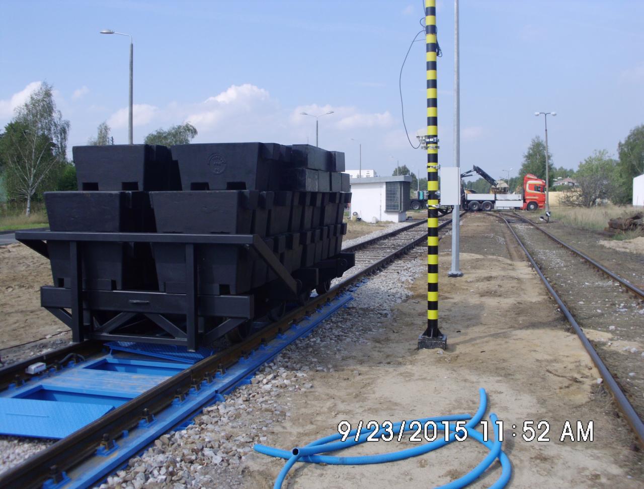 Wagonik z czarnymi blokami na torach, obok słup z czarno-żółtymi pasami. W tle dźwig na ciężarówce i budynek. Błękitny wąż na pierwszym planie.