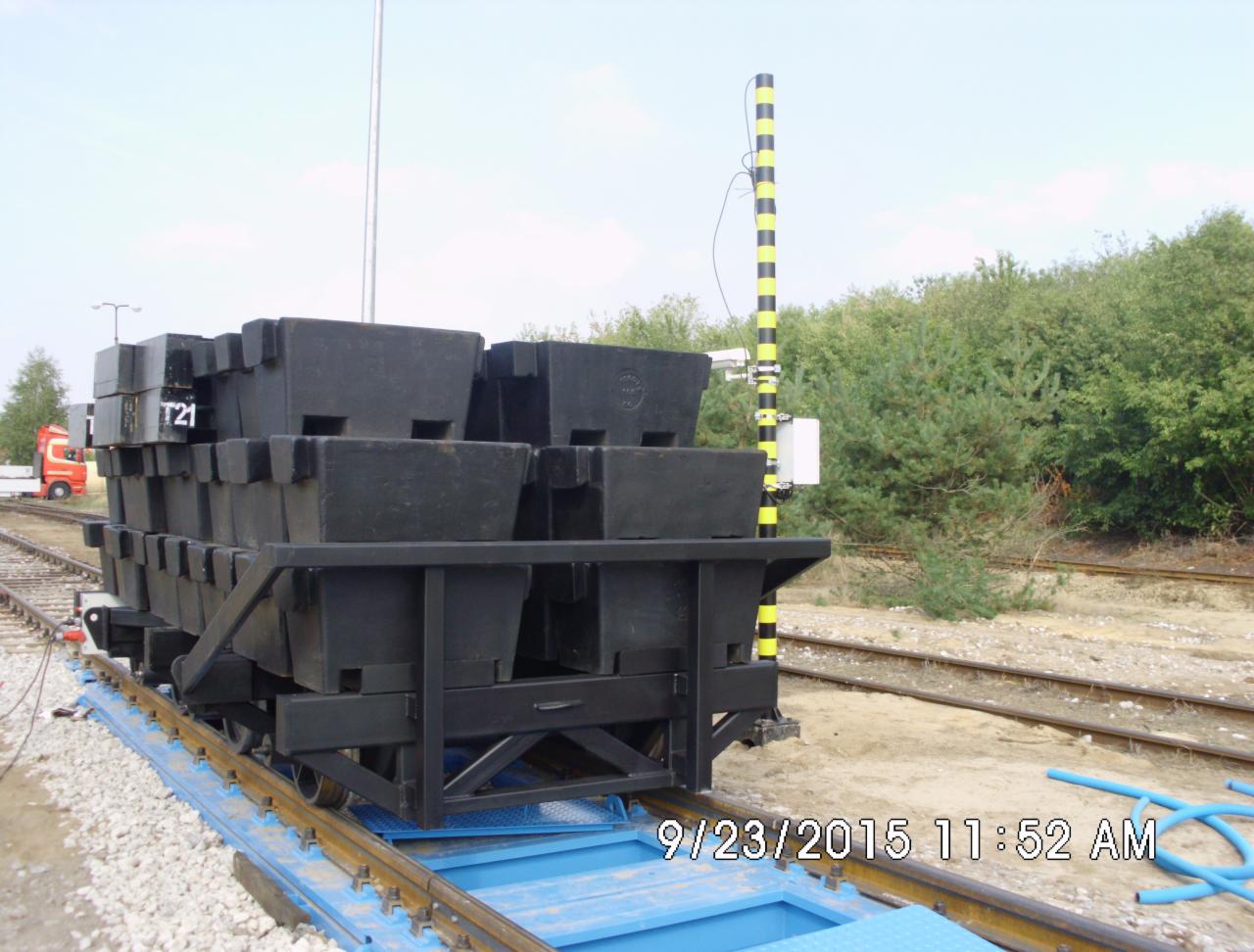 Wagonik kopalniany z czarnymi pojemnikami na szynach, widoczny z boku, z numerem T21. W tle zieleń i słup z żółto-czarnymi pasami. Data na dole zdjęcia.