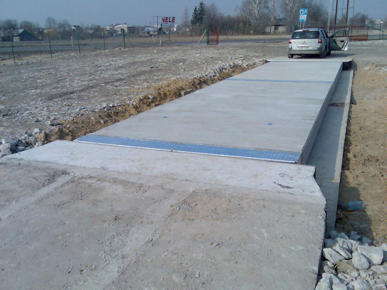 Betonowa waga samochodowa z najazdem, srebrnym wykończeniem i zaparkowanym obok autem z otwartymi drzwiami na placu budowy.