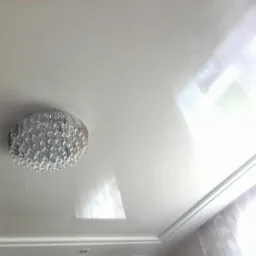 Biały, błyszczący sufit z zamontowaną, okrągłą lampą z kryształkami, widoczny fragment listwy przysufitowej i zasłony.