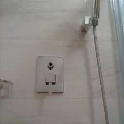 Wykończona łazienka: armatura chromowana, bateria podtynkowa i deszczownica na tle płytek imitujących marmur z dekoracyjnym pasem mozaiki u góry.