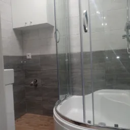 Narożna kabina prysznicowa z hydromasażem, przeszklona, z chromowanymi uchwytami, na tle szaro-białych płytek i szafki wiszącej w łazience z drewnopodobną podłogą.