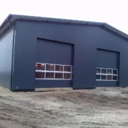 Nowoczesna hala z płyty warstwowej w kolorze grafitowym, z dwoma bramami segmentowymi z oknami. Teren wokół wyrównany piaskiem.