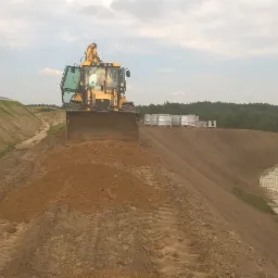 Żółta koparko-ładowarka wyrównuje teren pod budowę drogi, widoczne ułożone ażurowe płyty betonowe i składowane palety z kostką brukową.