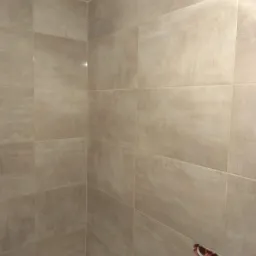 Beżowe, matowe płytki ceramiczne ułożone na ścianach w rogu łazienki nad białą wanną, widoczne gniazdko elektryczne w ścianie.