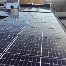 Montaż paneli fotowoltaicznych na płaskim dachu pokrytym papą, widoczny pracownik w pomarańczowym kombinezonie oraz narzędzia.