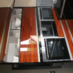 Kuchenne szafki z fornirem w odcieniach brązu i czerni, częściowo otwarte, ukazujące zawartość: naczynia i sprzęt AGD. Ujęcie z góry, blask lakieru.