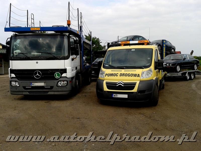 Dwa pojazdy pomocy drogowej: biały Mercedes Actros i żółty Citroen Jumper, z załadowanymi samochodami osobowymi na przyczepie. Widok z przodu, na szutrowym podłożu.