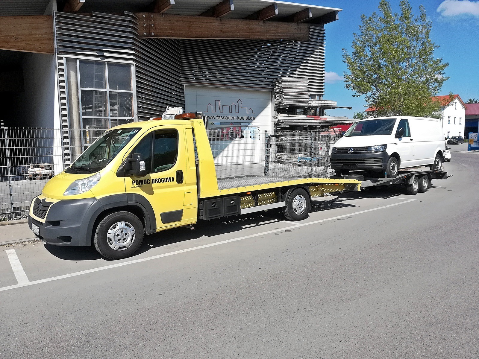 Żółta ciężarówka pomocy drogowej z białym vanem na platformie, zaparkowana na ulicy przed nowoczesnym budynkiem z żaluzjami.