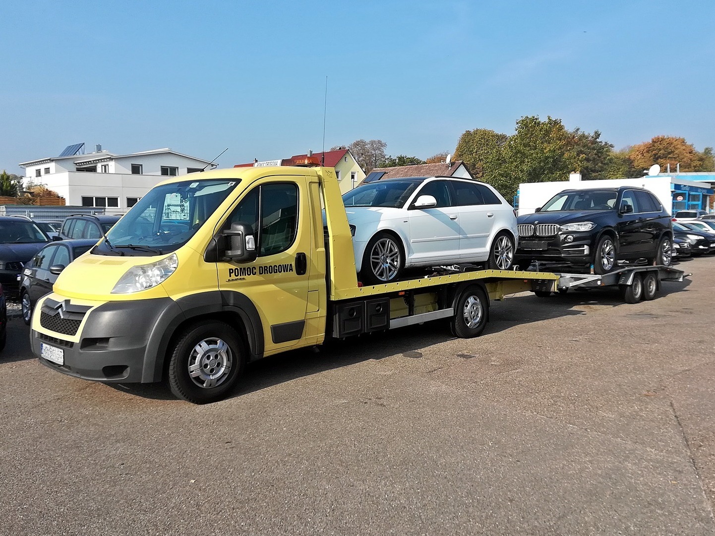 Żółta laweta Citroen z napisem 'Pomoc Drogowa' przewozi białe Audi A3 i czarne BMW X5 na parkingu