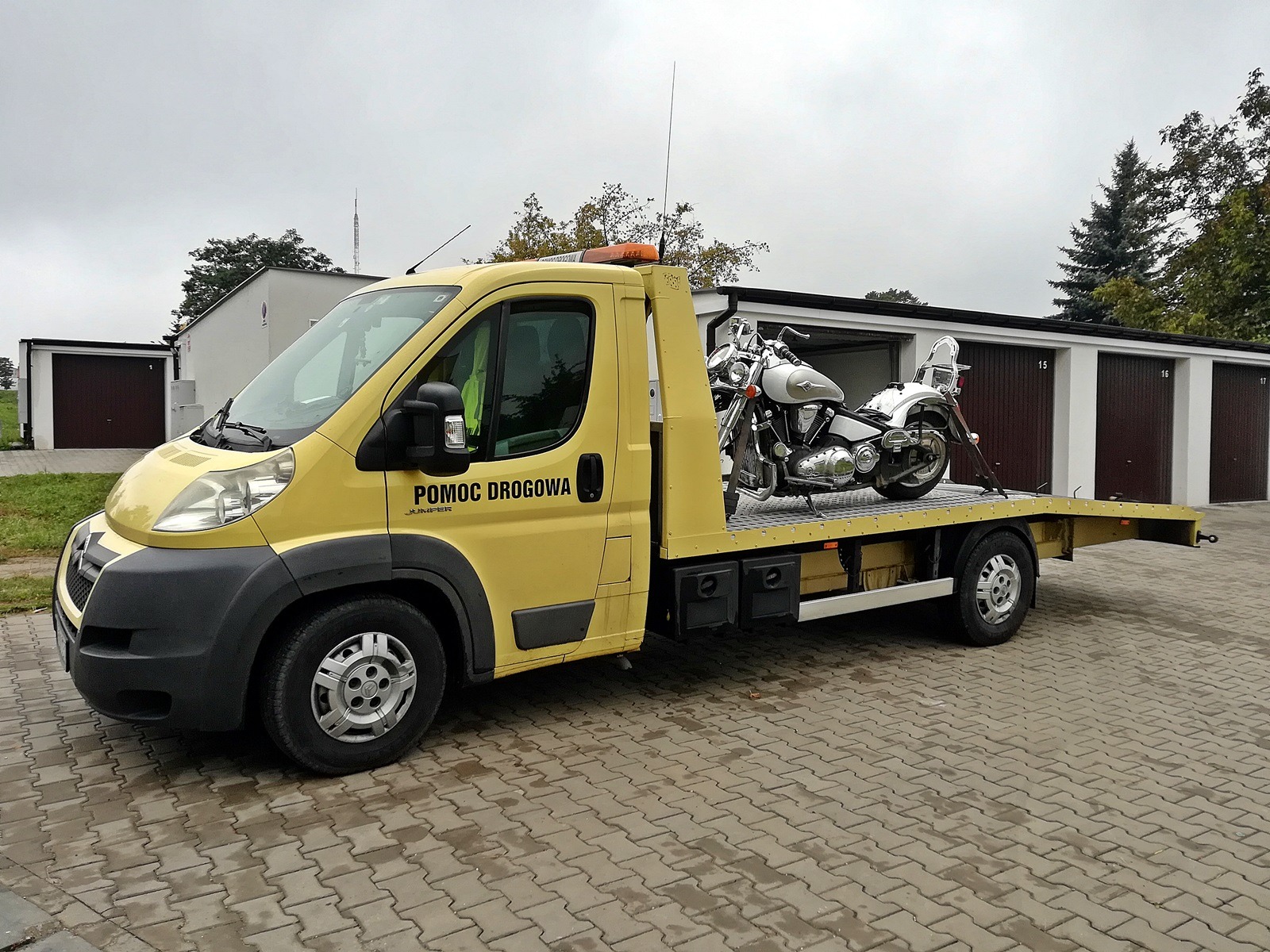Żółty pojazd pomocy drogowej z napisem 'POMOC DROGOWA' przewozi na platformie srebrny motocykl, na tle budynków garażowych.