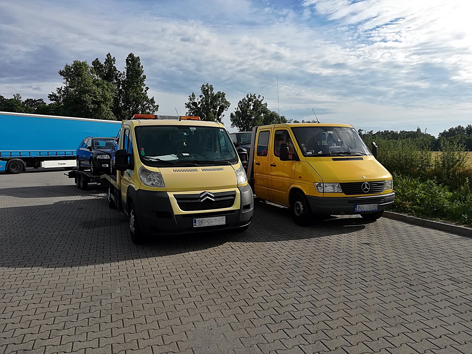 Żółta platforma transportowa marki Citroen z załadowanym niebieskim samochodem osobowym oraz żółty bus marki Mercedes-Benz na brukowanym placu postojowym w słoneczny dzień.