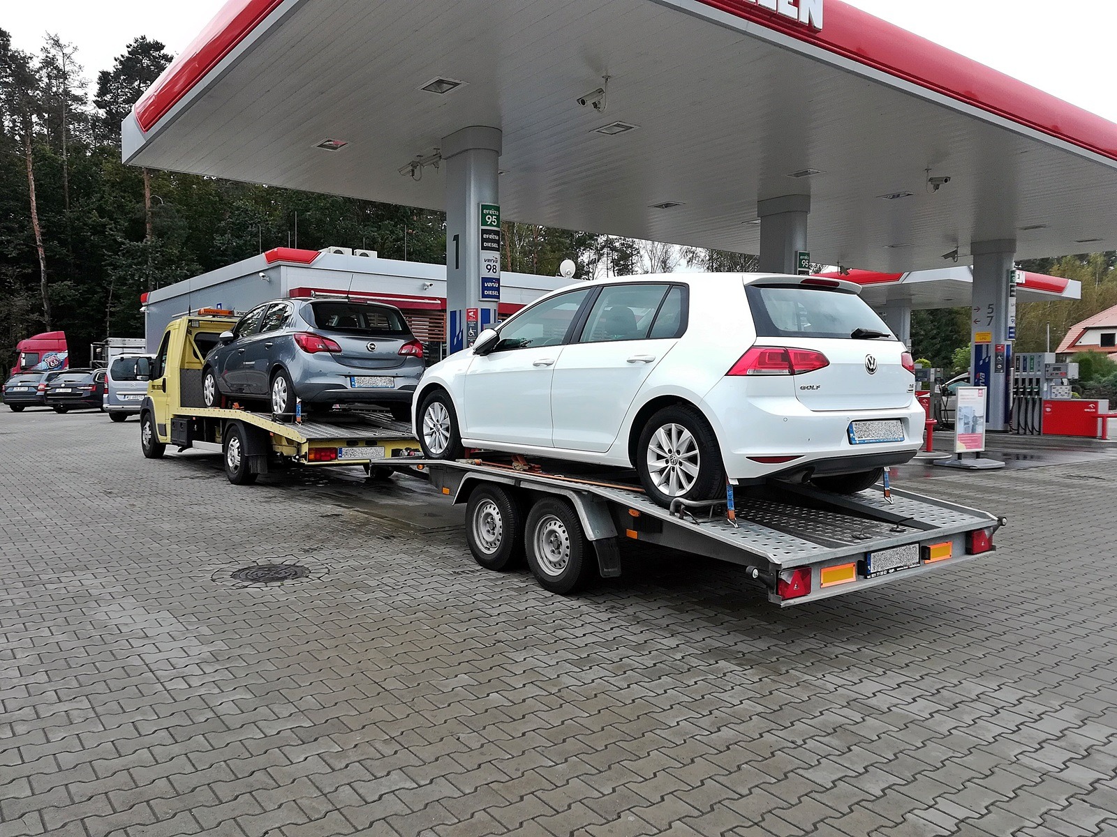 Żółty pojazd transportowy z dwoma samochodami osobowymi na platformie, zaparkowany na stacji benzynowej z widocznymi dystrybutorami i zadaszeniem.