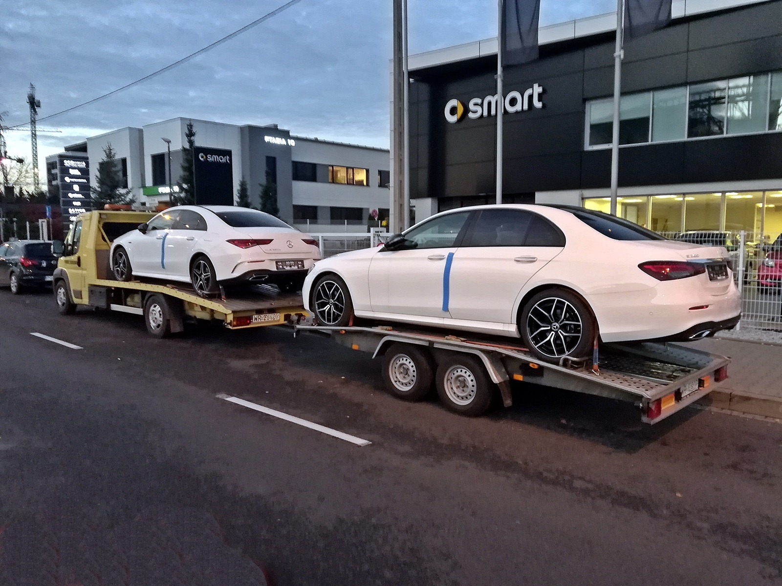 Żółta ciężarówka z dwoma białymi samochodami marki Mercedes na przyczepach, zaparkowana na ulicy przed salonem Smart.