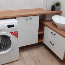 Biała pralka Electrolux obok szafki z blatem drewnianym i umywalką nablatową w nowoczesnej łazience z białymi ścianami.