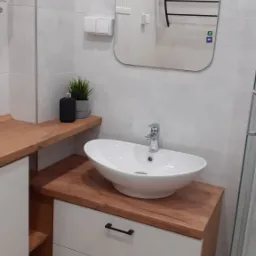 Biała umywalka nablatowa w kształcie łódki, zamontowana na szafce z dwiema szufladami z drewnianym blatem, uzupełniona czarnym dozownikiem mydła i małą rośliną doniczkową na półce, w tle lustro...