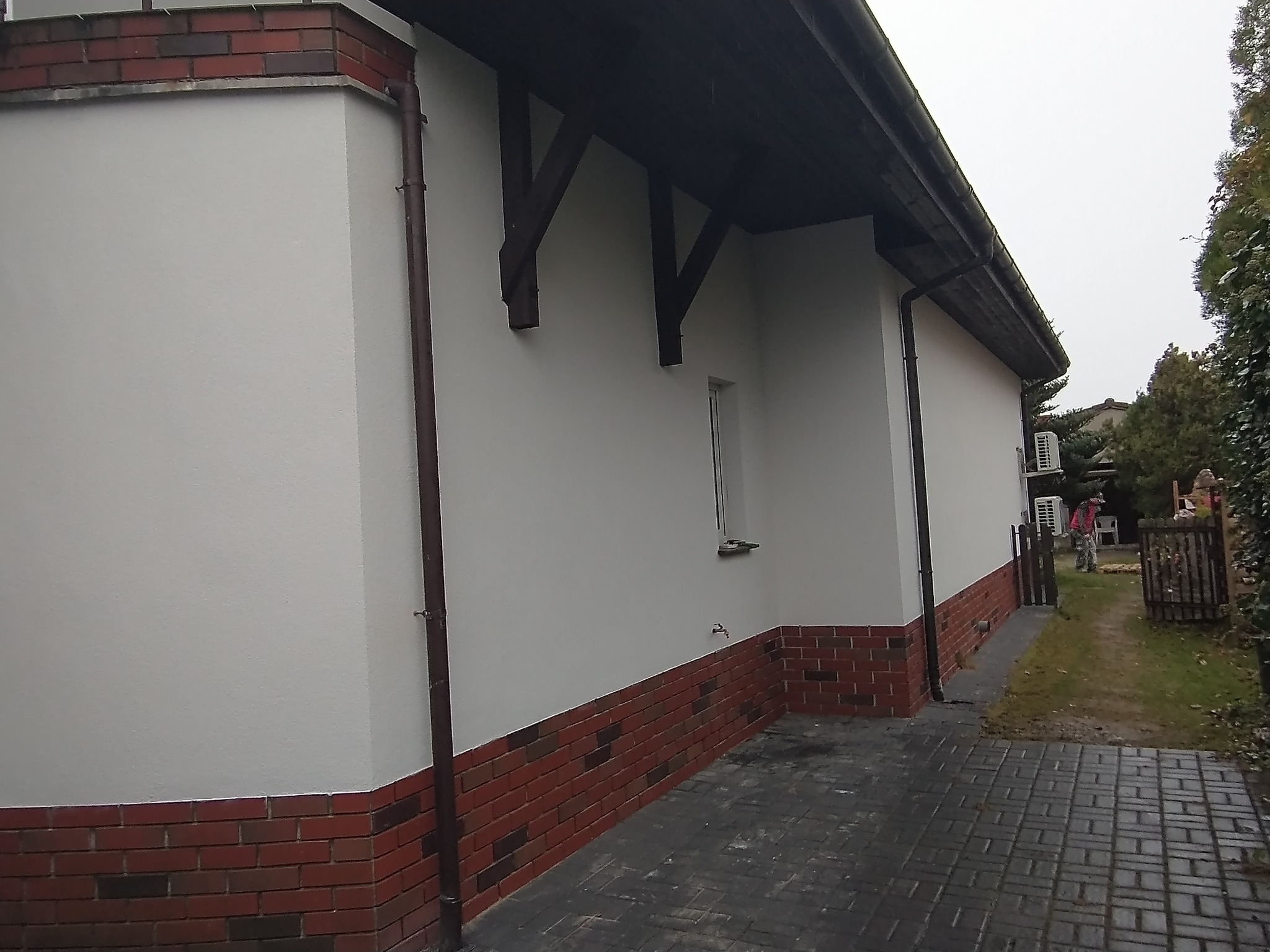 Elewacja budynku po remoncie: białe ściany z ceglaną podmurówką, brązowe rynny i drewniane wsporniki dachu, widoczny fragment chodnika z kostki brukowej.