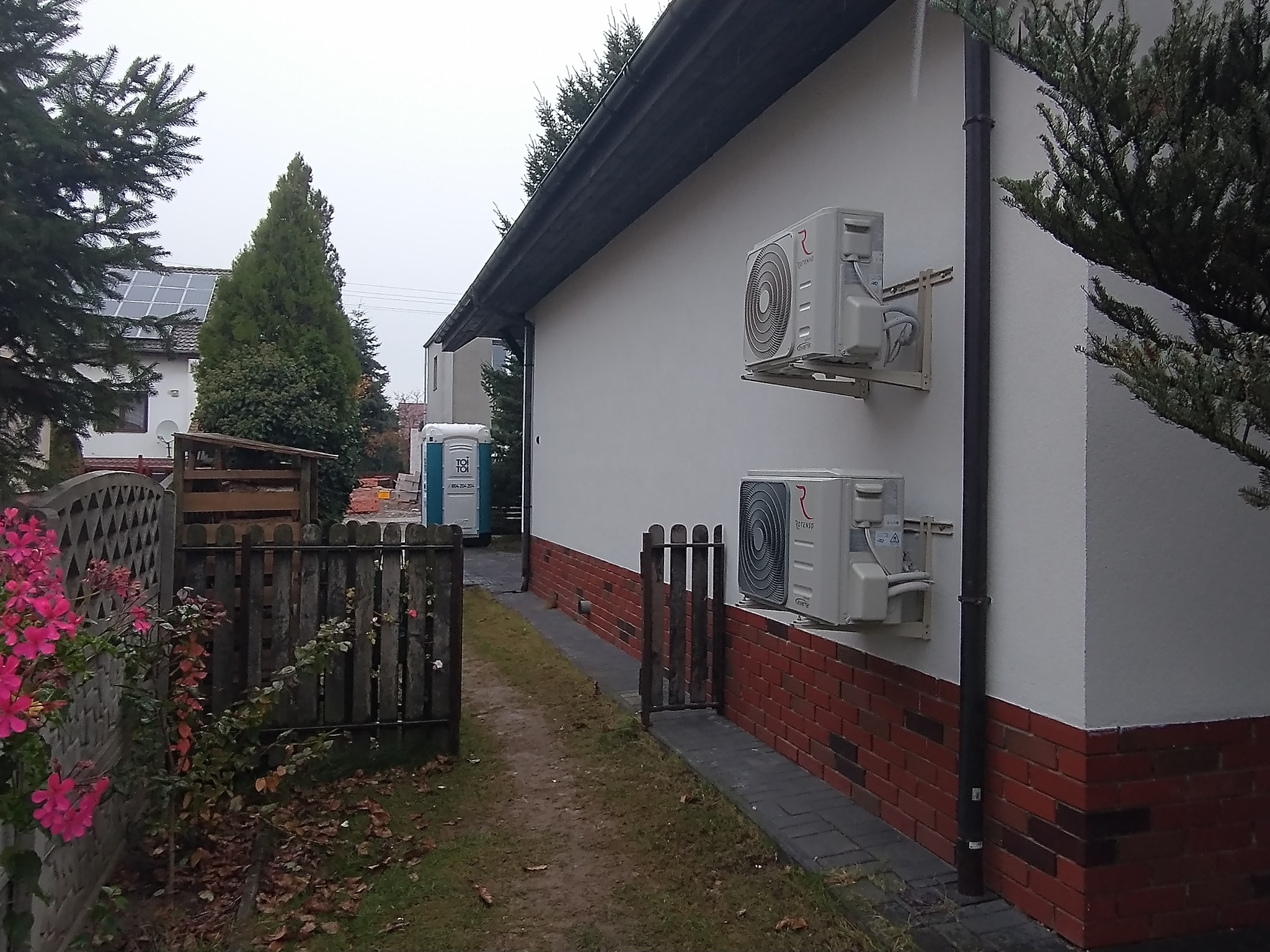 Dwa białe klimatyzatory zamontowane na zewnętrznej ścianie domu z czerwoną ceglaną podmurówką, widoczne z boku, w tle ogrodzenie i przenośna toaleta.
