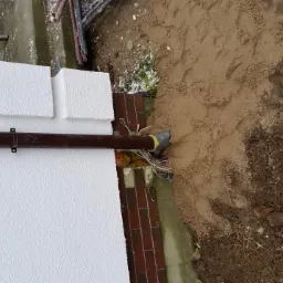 Biała elewacja z widoczną rurą spustową i fragmentem instalacji elektrycznej wystającej z ziemi. Widoczne cegły i piasek.