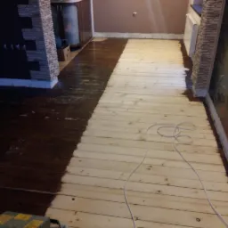 Częściowo polakierowana drewniana podłoga w trakcie renowacji w Żywcu, widoczny kabel i fragment ściany z dekoracyjnym kamieniem.