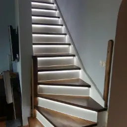 Drewniane schody z podświetleniem LED na każdym stopniu, widok z dołu, ciemne stopnie, białe podstopnice, drewniana barierka po prawej.