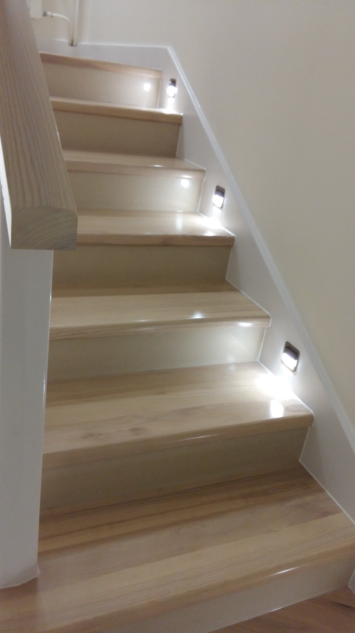 Jasne, drewniane schody z subtelnym oświetleniem LED w ścianie, widok z dołu. Naturalne wykończenie drewna i minimalistyczny design.