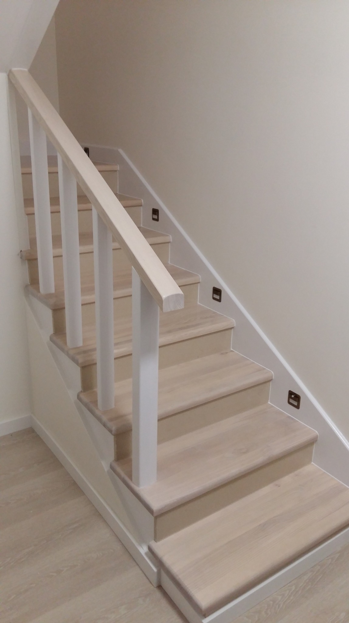 Jasne, drewniane schody z białymi balustradami i subtelnym oświetleniem punktowym w ścianie, tworzące minimalistyczny, nowoczesny design wnętrza.