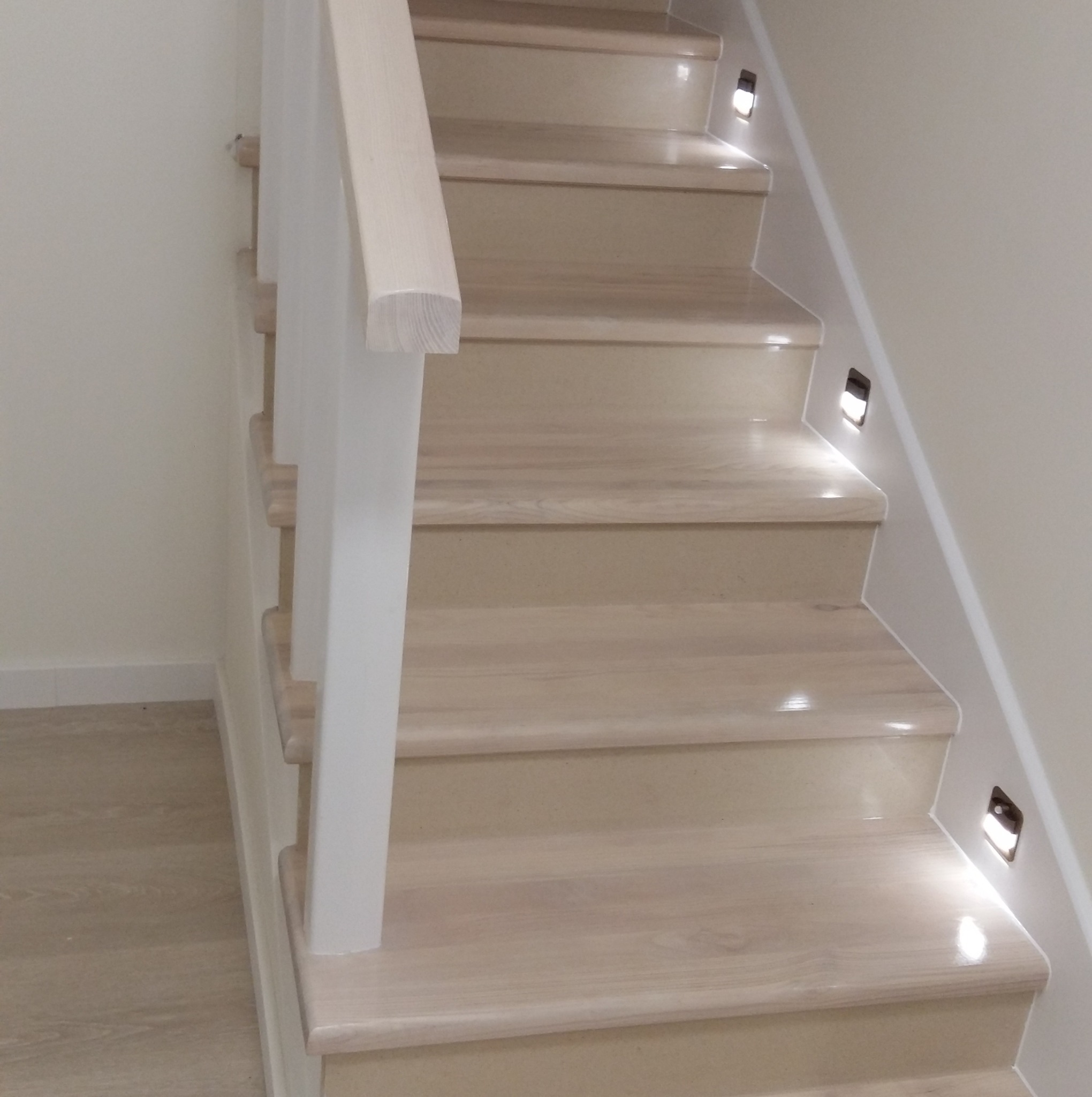 Jasne, drewniane schody z białymi balustradami i subtelnym oświetleniem LED wbudowanym w ścianę, tworzące nowoczesny i elegancki wygląd wnętrza.