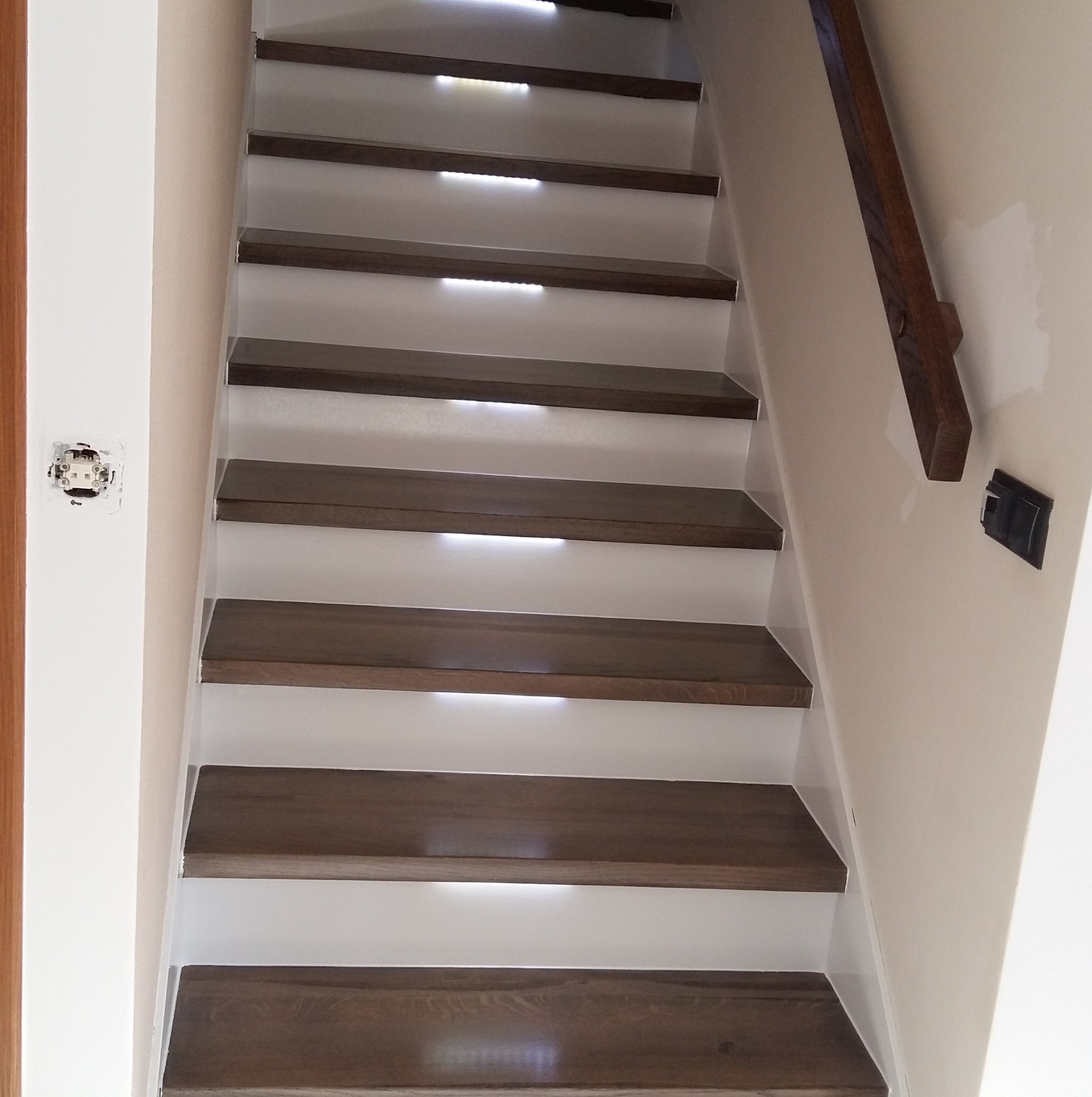 Nowoczesne schody drewniane z podświetleniem LED, brązowe stopnie i białe podstopnice. Widoczny fragment drewnianej poręczy i gniazdko elektryczne.