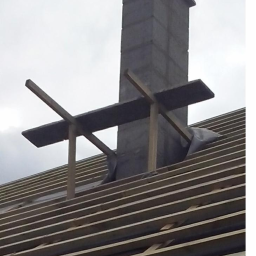 Dawwoj - Wznoszony komin z bloczków betonowych na drewnianej konstrukcji dachu z łatami, zabezpieczony folią. Surowy stan budowy, pochmurne niebo w tle.