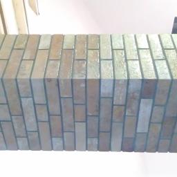 Dawwoj - Wykończony fragment ściany z cegiełkowych płytek w odcieniach beżu i brązu, widok z dołu, z elementami drewnianej konstrukcji sufitu w tle.