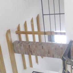 Dawwoj - Nowoczesna balustrada schodowa z drewnianymi elementami dekoracyjnymi na białej ścianie. Widok z dołu, fragment okna i kraty w tle.