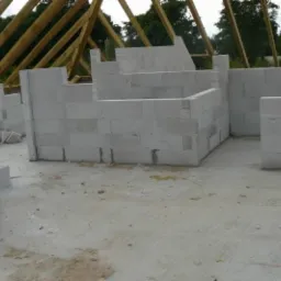 Wnętrze domu w trakcie budowy. Ściany z białych bloczków, drewniany szkielet dachu. Widoczne surowe wykończenie podłogi i ściany w trakcie wznoszenia.