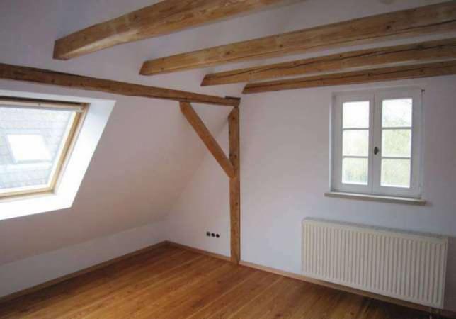 Jasne poddasze z drewnianymi belkami stropowymi i podłogą. Okno dachowe w skosie, białe ściany i grzejnik. Widoczne elementy konstrukcyjne z drewna.