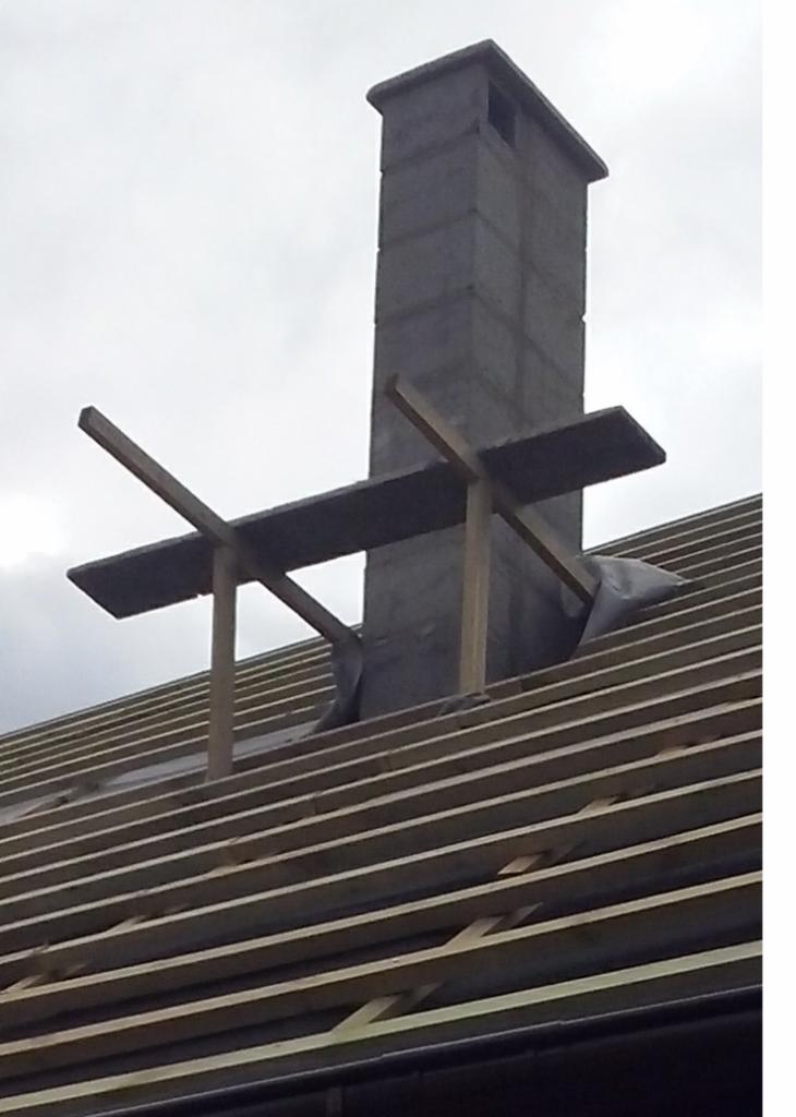 Wznoszony komin z bloczków betonowych na drewnianej konstrukcji dachu z łatami, zabezpieczony folią. Surowy stan budowy, pochmurne niebo w tle.