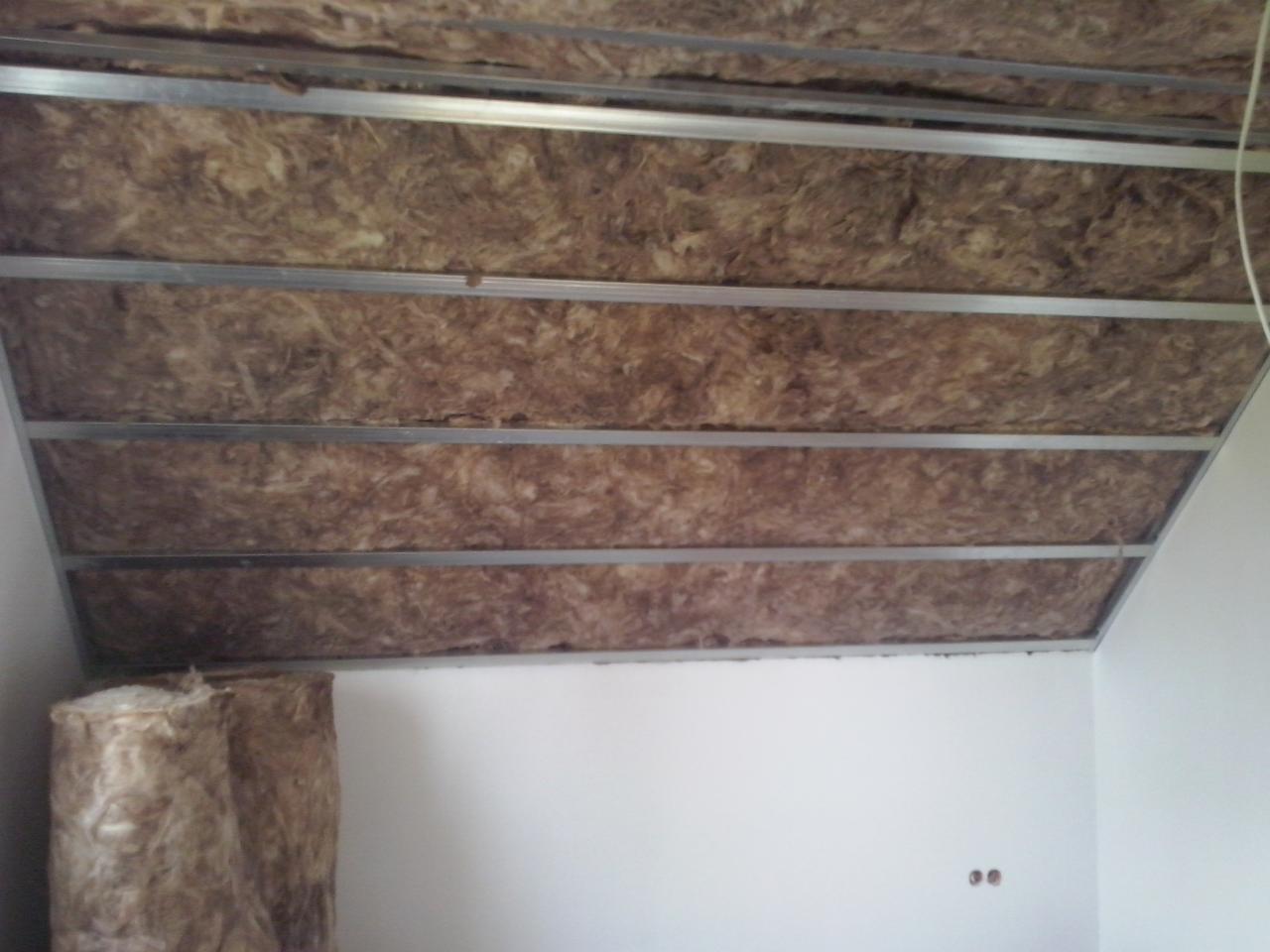 Izolacja poddasza wełną mineralną z widoczną metalową konstrukcją sufitu podwieszanego i rolką wełny w rogu. Białe ściany, widoczne gniazdka elektryczne.