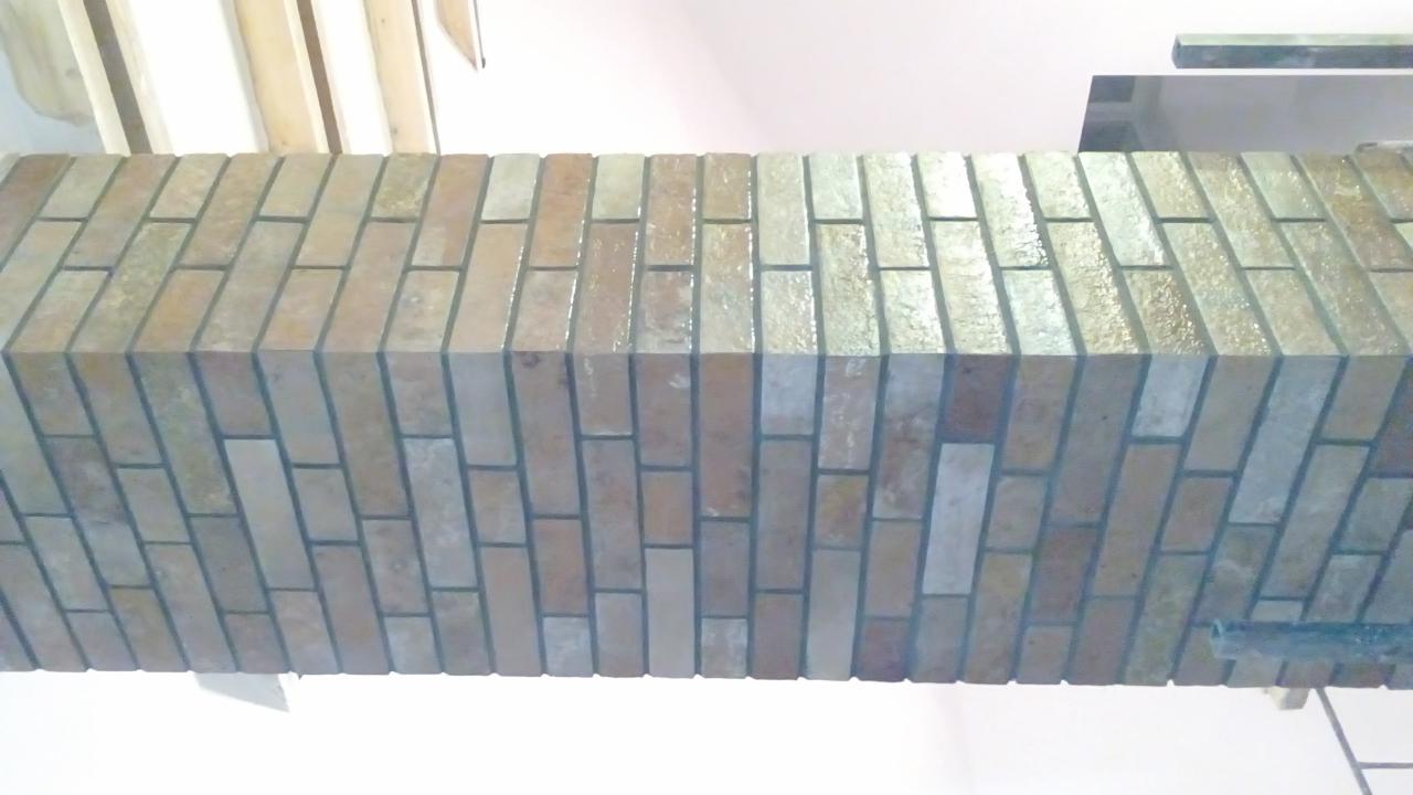 Wykończony fragment ściany z cegiełkowych płytek w odcieniach beżu i brązu, widok z dołu, z elementami drewnianej konstrukcji sufitu w tle.