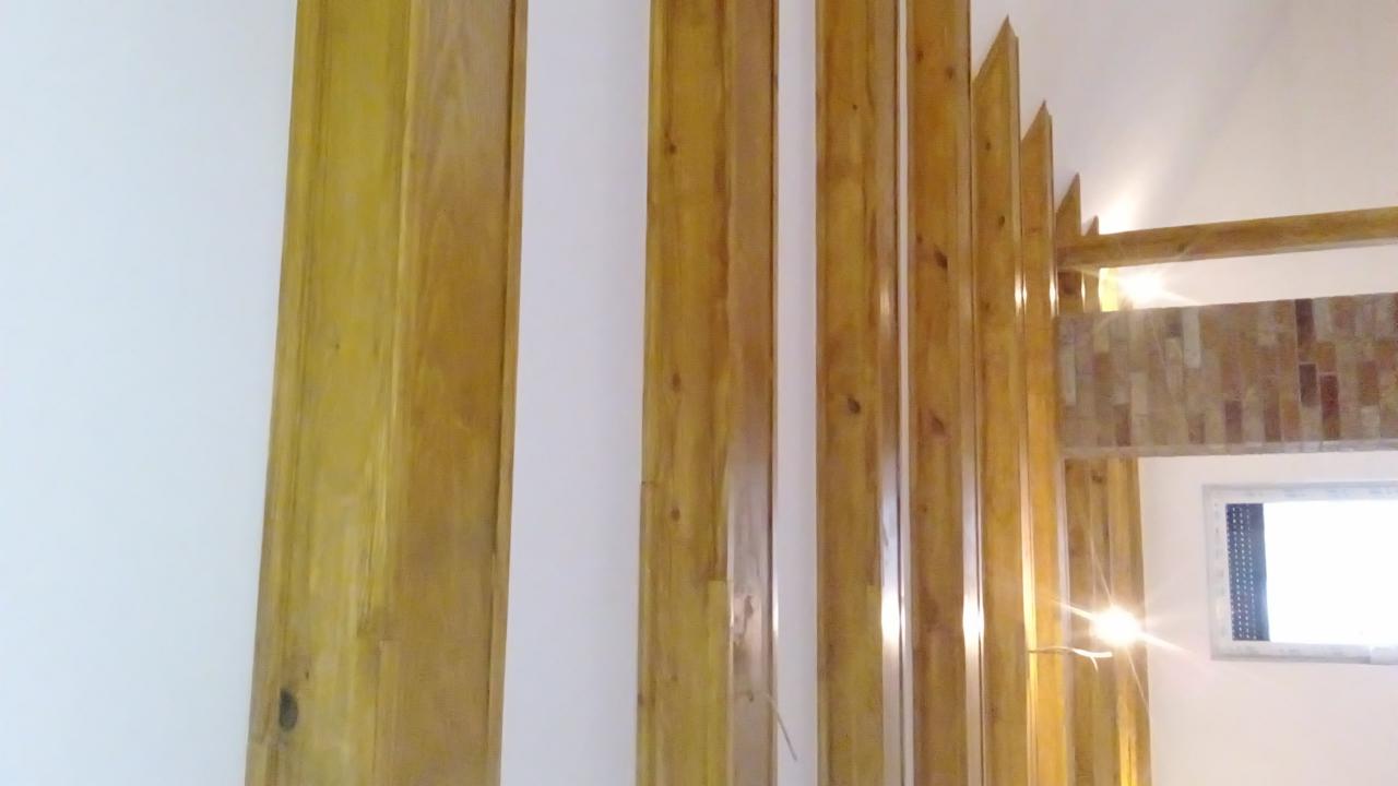 Drewniane lamele pionowe, tworzące ażurową ściankę działową, z widocznym fragmentem okna w tle. Naturalny kolor drewna, jasne, neutralne tło.