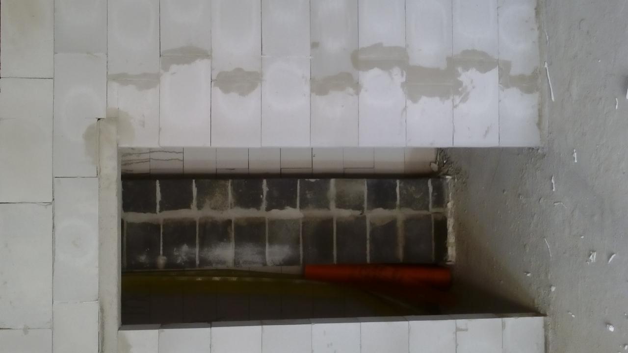 Wnętrze w trakcie budowy: białe bloczki ścienne, widoczna instalacja rur w otworze, surowy beton na podłodze. Etap prac instalacyjnych przed dalszym wykończeniem.