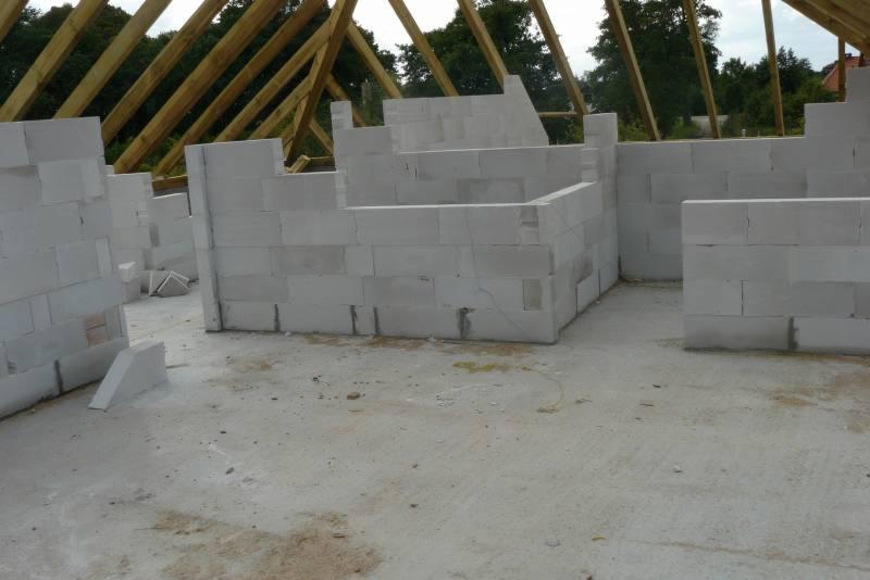 Wnętrze domu w trakcie budowy. Ściany z białych bloczków, drewniany szkielet dachu. Widoczne surowe wykończenie podłogi i ściany w trakcie wznoszenia.