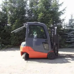 Nowy wózek widłowy Toyota z otwartą kabiną, pomarańczowo-szary, na żwirowym podłożu, w tle iglaki i opony.